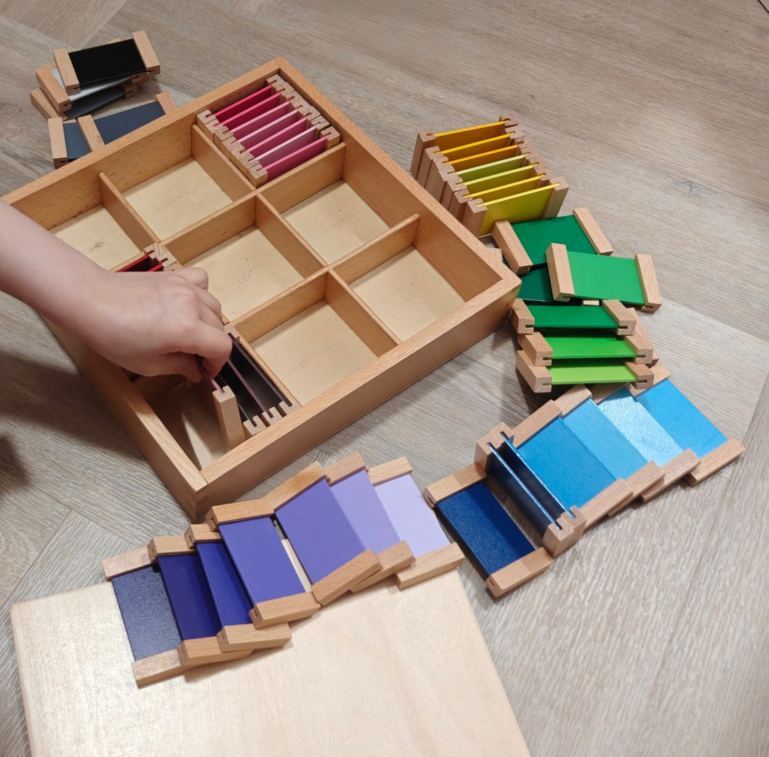 A child’s hand carefully arranging colourful wooden Montessori Color Tablets in a divided wooden sorting box on a light herringbone floor.